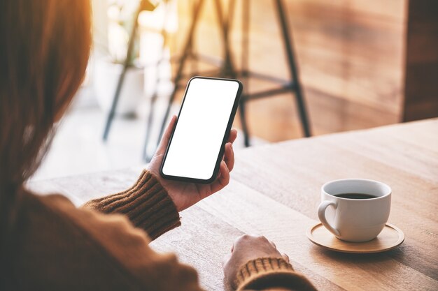 Immagine mockup di una donna che tiene in mano un telefono cellulare nero con uno schermo desktop vuoto con una tazza di caffè sul tavolo