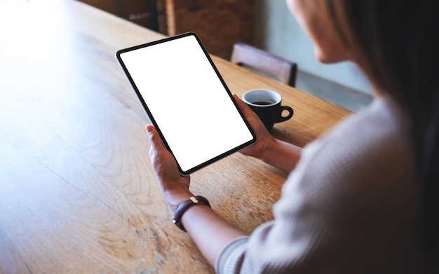 Immagine mockup di una donna che tiene tablet digitale con schermo desktop bianco vuoto con tazza di caffè sul tavolo di legno