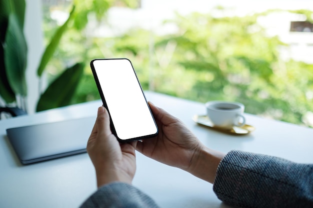 Immagine mockup di una donna d'affari che tiene in mano un telefono cellulare con uno schermo desktop bianco vuoto con un laptop sul tavolo