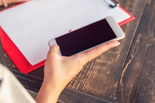 Immagine mockup di una mano che tiene un telefono cellulare bianco con schermo desktop nero vuoto sullo sfondo di un foglio di carta bianco in un bar.