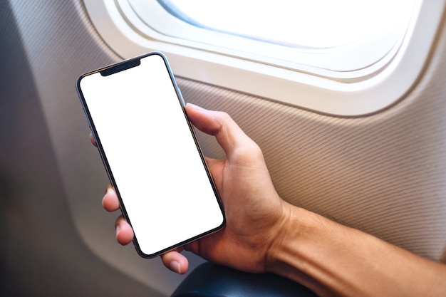 Immagine mockup di una mano che tiene un telefono cellulare nero con schermo desktop vuoto accanto a una finestra dell'aeroplano