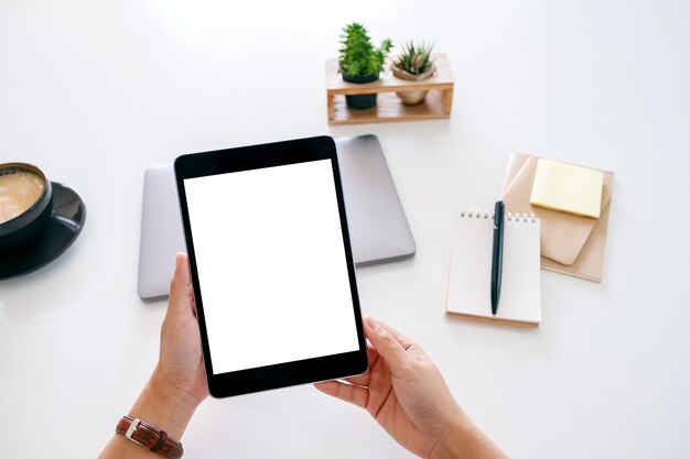 Immagine mockup vista dall'alto delle mani che tengono un tablet con schermo bianco vuoto e computer portatile su tavolo di legno in ufficio