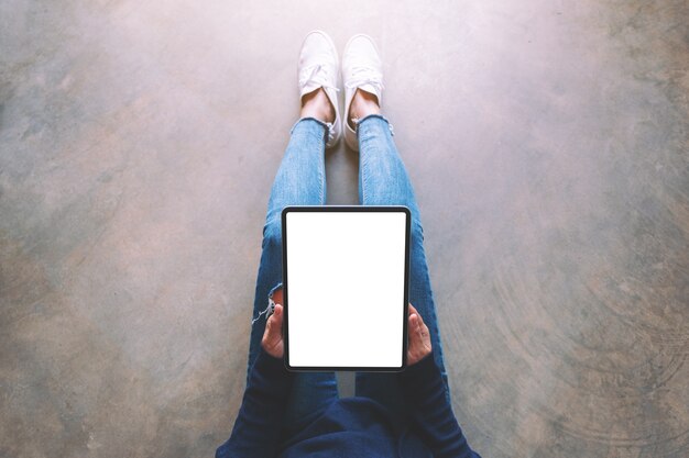 Immagine mockup vista dall'alto di una donna che tiene in mano un tablet pc nero con schermo bianco vuoto mentre è seduta sul pavimento