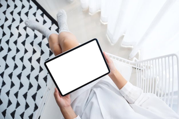 Immagine mockup vista dall'alto di una donna che tiene in mano un tablet pc nero con uno schermo desktop bianco vuoto mentre è seduta in camera da letto con una sensazione di relax al mattino