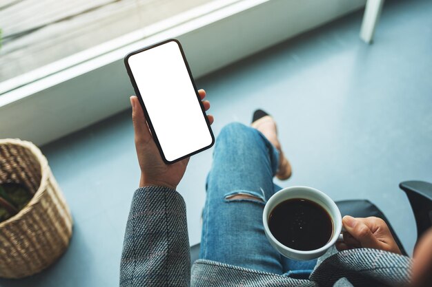 Immagine mockup vista dall'alto di una donna che tiene in mano un telefono cellulare con uno schermo desktop bianco vuoto con una tazza di caffè