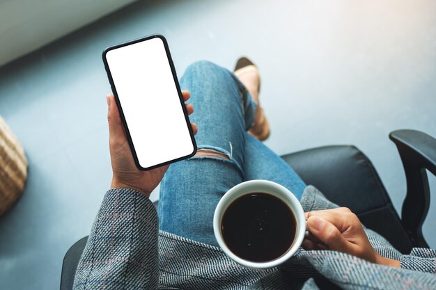 Immagine mockup vista dall'alto di una donna che tiene in mano un telefono cellulare con uno schermo desktop bianco vuoto con una tazza di caffè