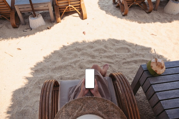 Immagine mockup vista dall'alto di una donna che tiene in mano un telefono cellulare con uno schermo desktop vuoto mentre è seduto sulla spiaggia