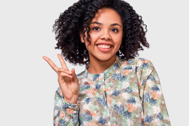 Immagine orizzontale del primo piano della giovane donna afroamericana positiva che sorride ampiamente mostrando il gesto di pace mentre guarda alla macchina fotografica che sta contro il fondo bianco dello studio