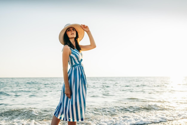 Immagine orizzontale della giovane donna castana attraente con il cappello bianco sulla testa che cammina sulla spiaggia e sul mare sullo sfondo del tramonto Bella donna che indossa un abito a righe godersi la brezza dell'oceano