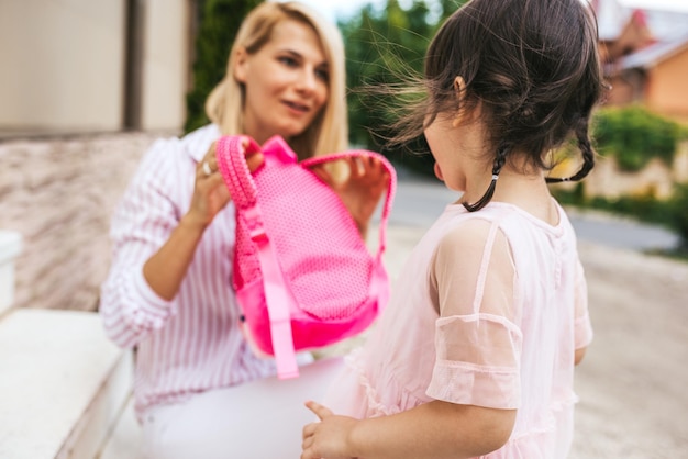 Immagine orizzontale di felice madre stupita che prepara lo zaino da mettere sullo zaino a suo figlio per andare all'asilo all'aperto Bambina carina mostra la lingua a sua madre prima del primo giorno di scuola materna