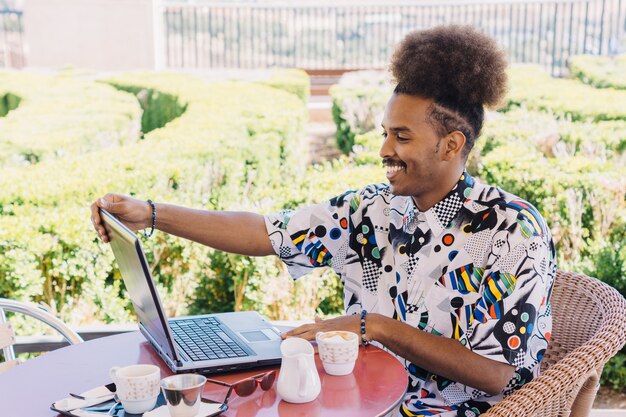 Immagine orizzontale di un ragazzo nero con i capelli afro raccolti sorridente seduto su una terrazza che lavora con il computer portatile a un tavolo rosso rotondo con tazze di caffè e un orto verde sfocato