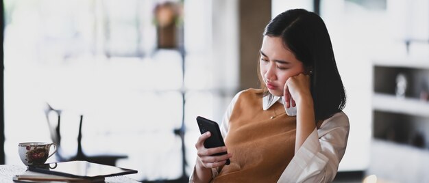 Immagine orizzontale di una giovane donna accigliata e frustrata che guarda lo smartphone sentendosi insoddisfatta dell'e-mail ricevuta.