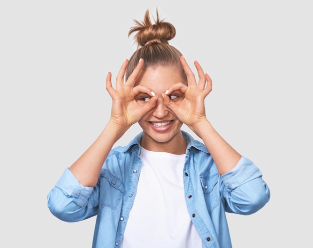 Immagine orizzontale di una giovane donna felice con un'acconciatura da panino bionda che mostra gesti ok con entrambe le mani che fingono di indossare occhiali o un binocolo Bella donna sorridente ampiamente e in posa sul muro bianco