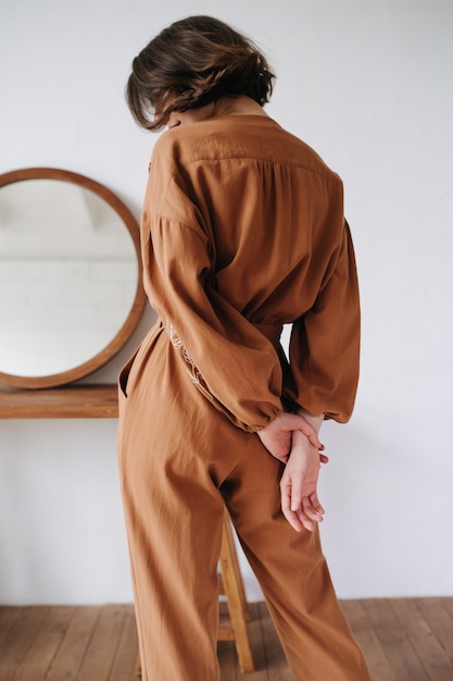 Immagine posteriore di una donna sexy in una tuta marrone che si tiene per mano dietro la schiena, sbirciando tra le ciocche di capelli. Bella bruna con i capelli corti in casa, accanto a un tavolo con uno specchio rotondo