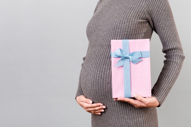 Immagine potata del contenitore di regalo macchiato rosa in mano della donna incinta contro la sua pancia a fondo grigio. Futura madre in abito grigio. In attesa di una bambina. Copia spazio