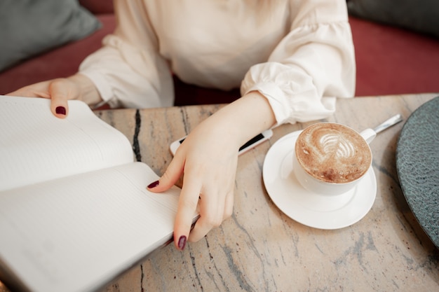 Immagine potata della ragazza che fa le note in blocco note aperto mentre sedendosi alla tavola e riposando in caffè accogliente moderno