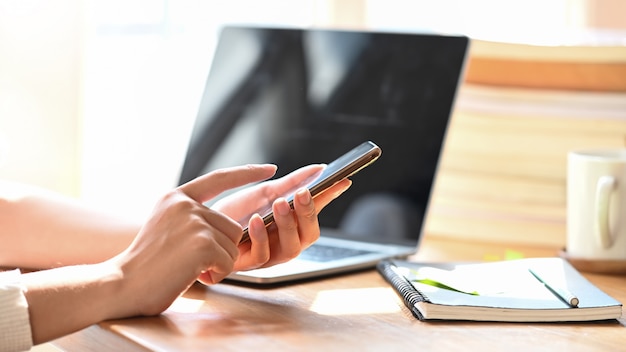 Immagine potata delle mani della donna che tengono smartphone allo scrittorio di legno.