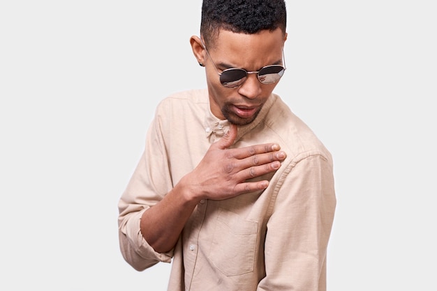 Immagine ravvicinata del giovane afroamericano che indossa occhiali da sole rotondi a specchio e camicia beige casual che fa un gesto swag e posa su sfondo bianco per studio Concetto di emozione della gente