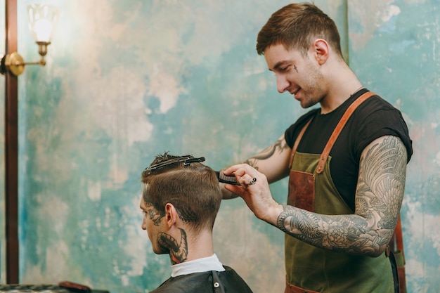 Immagine ravvicinata dell'uomo che ottiene taglio di capelli alla moda al negozio di barbiere. L'hairstylist maschile in tatuaggi al servizio del cliente.