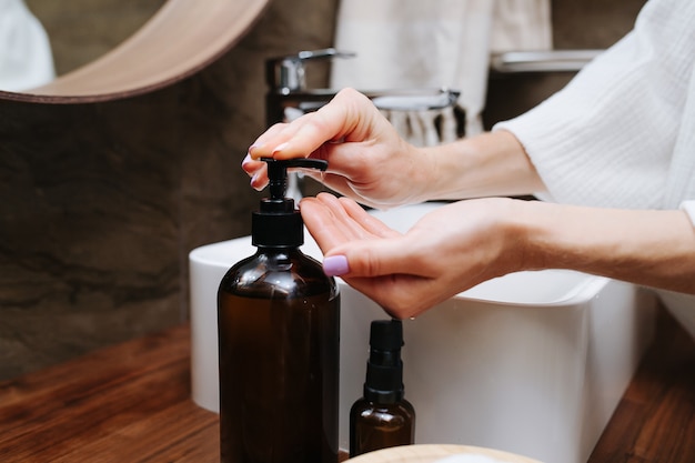 Immagine ravvicinata delle mani di una donna matura che pompa un sapone ecologico da una bottiglia. È in piedi davanti al lavandino in un bagno.