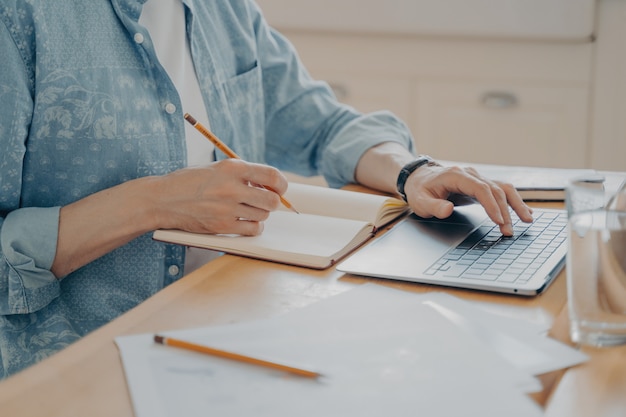 Immagine ravvicinata di mani maschili che digitano sul computer portatile per scrivere un nuovo articolo nel sito Web e prendere appunti in agenda al tavolo di legno della cucina. Concetto di freelance e lavoro a distanza da casa