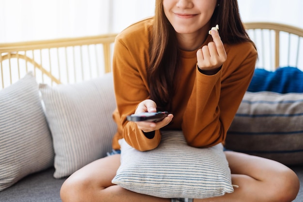 Immagine ravvicinata di una bellissima giovane donna che mangia pop corn e cerca un canale con il telecomando per guardare la tv mentre è seduta sul divano di casa