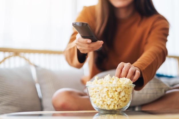 Immagine ravvicinata di una giovane donna che mangia popcorn e cerca un canale con il telecomando per guardare la tv mentre è seduta sul divano di casa