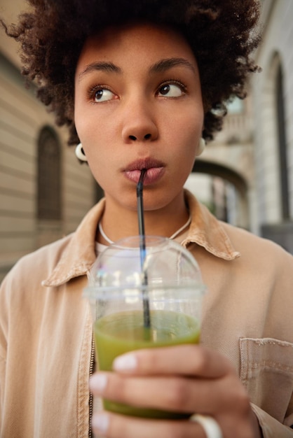 Immagine ravvicinata di una ragazza adolescente riccia pensierosa beve succo verde contenente molte vitamine pensa a uno stile di vita sano vestito con una giacca beige concentrata lontano Dieta disintossicante e concetto di nutrizione