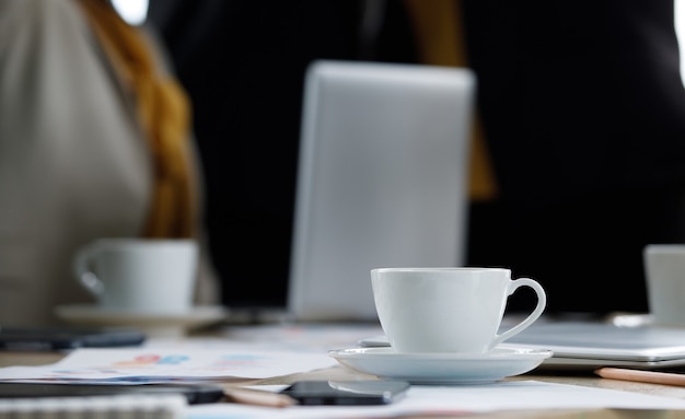 Immagine ravvicinata di una tazza di caffè caldo bianco e di un piccolo piatto sul tavolo della riunione vicino alla matita mentre un uomo d'affari irriconoscibile non identificato si prende una breve pausa dopo aver discusso e pianificato in uno sfondo sfocato.