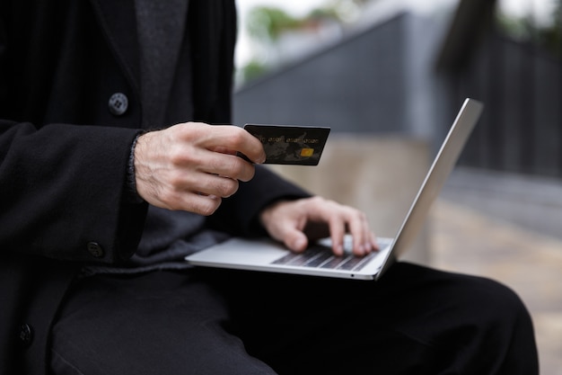 Immagine ritagliata del bel giovane imprenditore seduto all'aperto utilizzando il computer portatile in possesso di carta di credito.