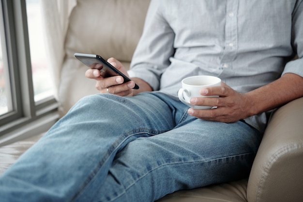 Immagine ritagliata dell'uomo seduto in poltrona che beve una tazza di caffè e controlla i social media sullo smartphone