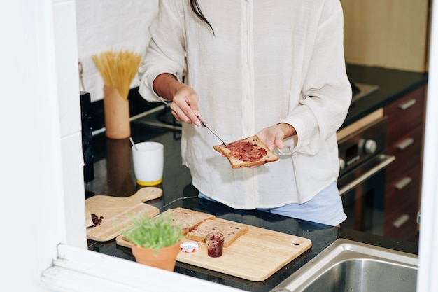 Immagine ritagliata della giovane donna che fa panino con gelatina per colazione utilizzando pane integrale