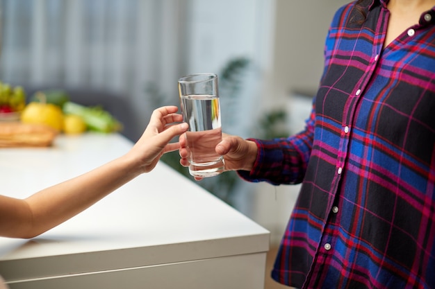 Immagine ritagliata della mano di una bambina che tiene un bicchiere d'acqua in cucina con la giovane madre.