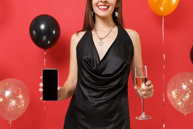 Immagine ritagliata della ragazza in abito nero che tiene in mano un bicchiere di champagne, telefono cellulare con schermo vuoto nero vuoto su mongolfiere di sfondo rosso brillante. Felice Anno Nuovo compleanno mockup festa concetto.