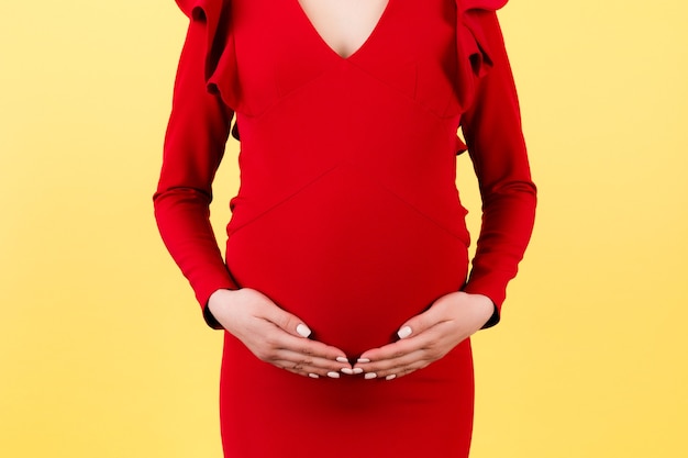 Immagine ritagliata delle mani della donna incinta che abbraccia il suo addome su sfondo giallo. La futura mamma indossa un vestito rosso. In attesa di un bambino. Copia spazio.