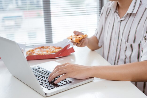 Immagine ritagliata di un uomo asiatico che pranza con la pizza mentre lavora alla sua scrivania.