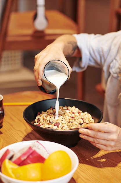 Immagine ritagliata di una donna che versa un bicchiere di latte in una ciotola di muesli con bacche secche o uvetta