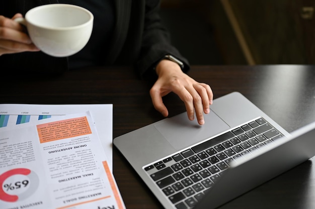 Immagine ritagliata di una donna d'affari che utilizza il suo laptop mentre sorseggia un caffè alla sua scrivania