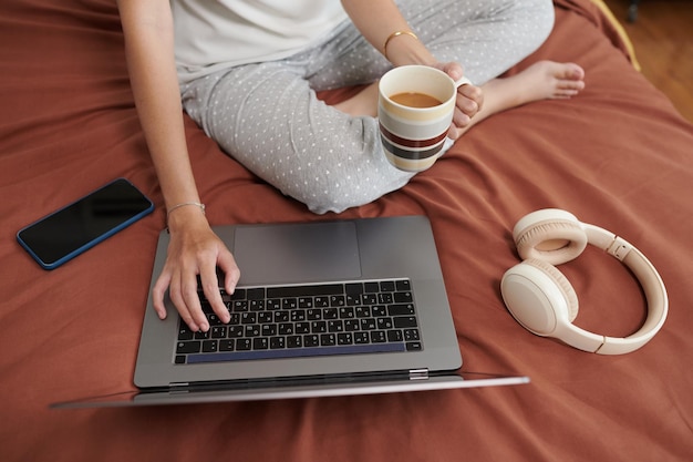 Immagine ritagliata di una donna in pigiama seduta sul letto a bere caffè e controllare la posta elettronica