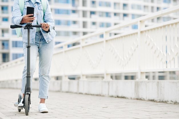 Immagine ritagliata di una giovane donna in jeans in piedi su uno scooter elettrico con lo smartphone in mano