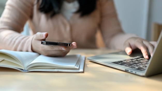 Immagine ritagliata di una giovane studentessa universitaria asiatica che tiene il suo telefono e usa il suo laptop