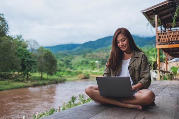 Immagine ritratto di una bella donna asiatica che lavora al computer portatile mentre è seduta in riva al fiume con montagne e sfondo naturale