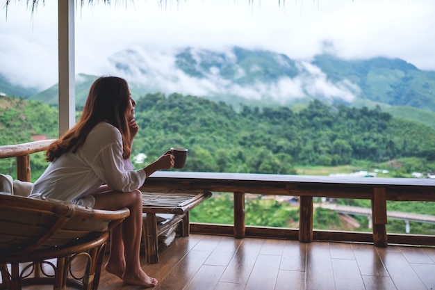 Immagine ritratto di una bella donna asiatica che tiene e beve caffè caldo sul balcone, guardando le montagne e la natura verde