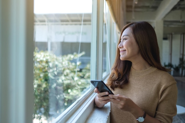 Immagine ritratto di una bella giovane donna asiatica che guarda fuori dalla finestra mentre si tiene e si utilizza il telefono cellulare