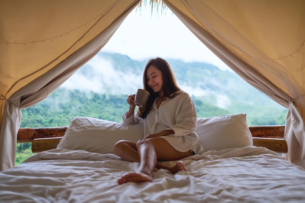 Immagine ritratto di una bellissima giovane donna asiatica che beve caffè mentre è seduta su un letto bianco al mattino con una bellissima vista sulla natura fuori dalla tenda