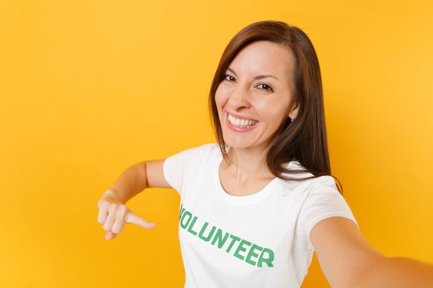 Immagine selfie di donna soddisfatta sorridente felice in maglietta bianca con iscrizione scritta volontario titolo verde isolato su sfondo giallo. Aiuto volontario di assistenza gratuita, concetto di lavoro di grazia di carità.