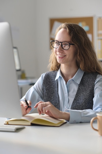 Immagine verticale di una giovane donna sorridente che usa il computer al lavoro mentre è seduta al suo posto di lavoro con