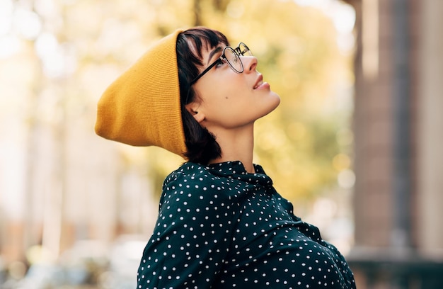 Immagine vista laterale di una bella giovane donna che indossa un cappello giallo e merda verde con punti bianchi che guarda in alto e sogna Giovane studentessa universitaria che si rilassa nella strada della città