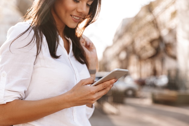 Immagini ritagliate di donna bruna soddisfatta in camicia utilizzando lo smartphone