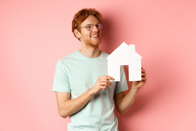 Immobiliare. Uomo rosso felice che sogna di acquistare proprietà, guardando a destra e mostrando il ritaglio di casa di carta, in piedi su sfondo rosa.